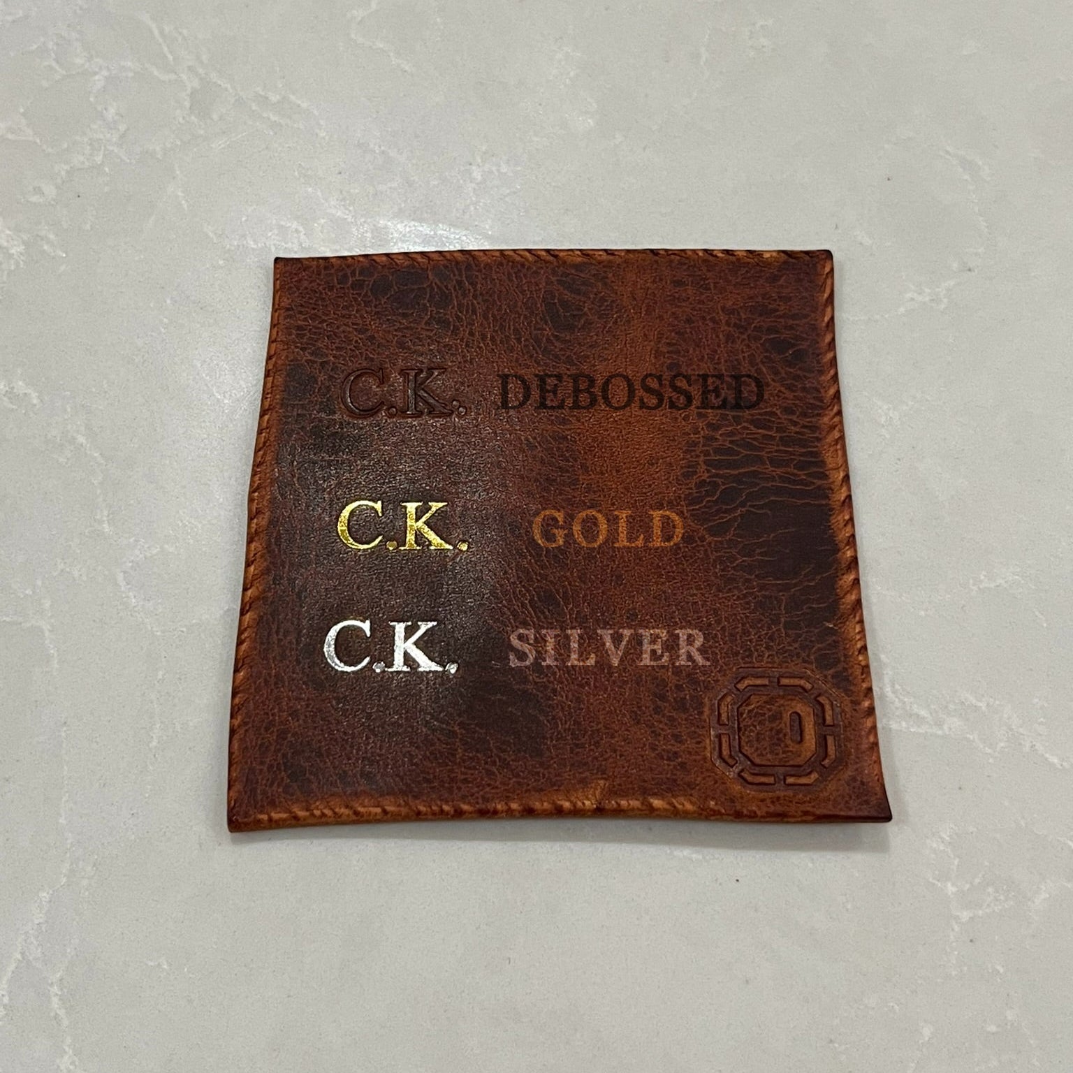 Brown leather patch with debossed and foiled text on a light gray background.
Displaying how gold and silver foiled and debossed monogramming appears on a patch of leather