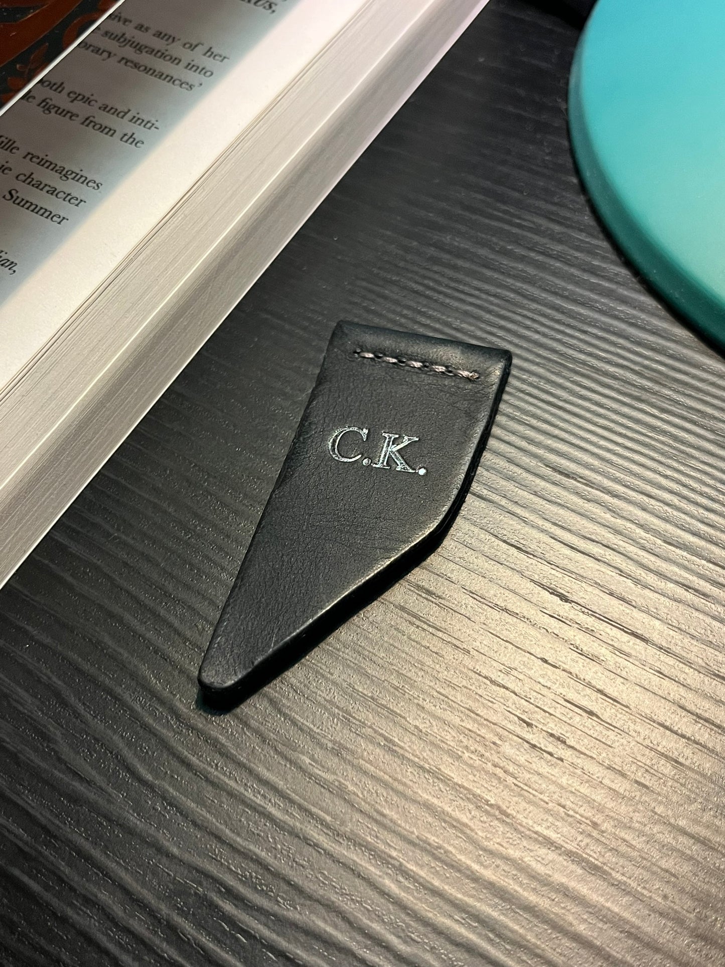 Dark navy leather bookmark with initials 'C.K.' with silver foil on a textured surface next to an open book.