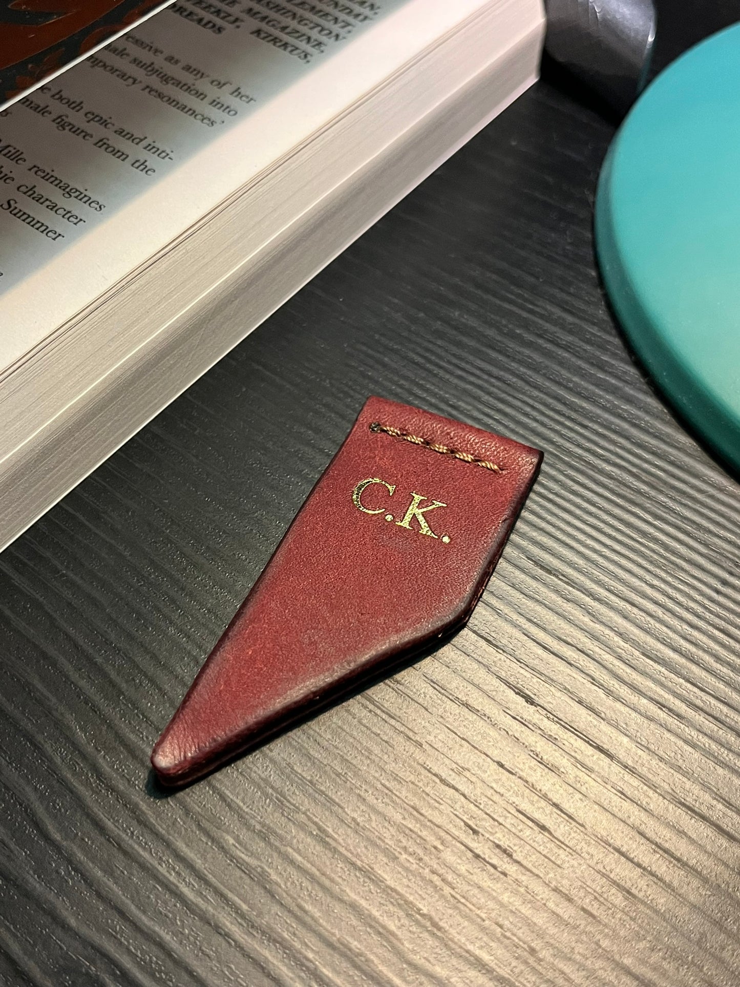 Red ribbon-style leather bookmark with initials 'C.K.' with gold foil on a wooden surface next to an open book.