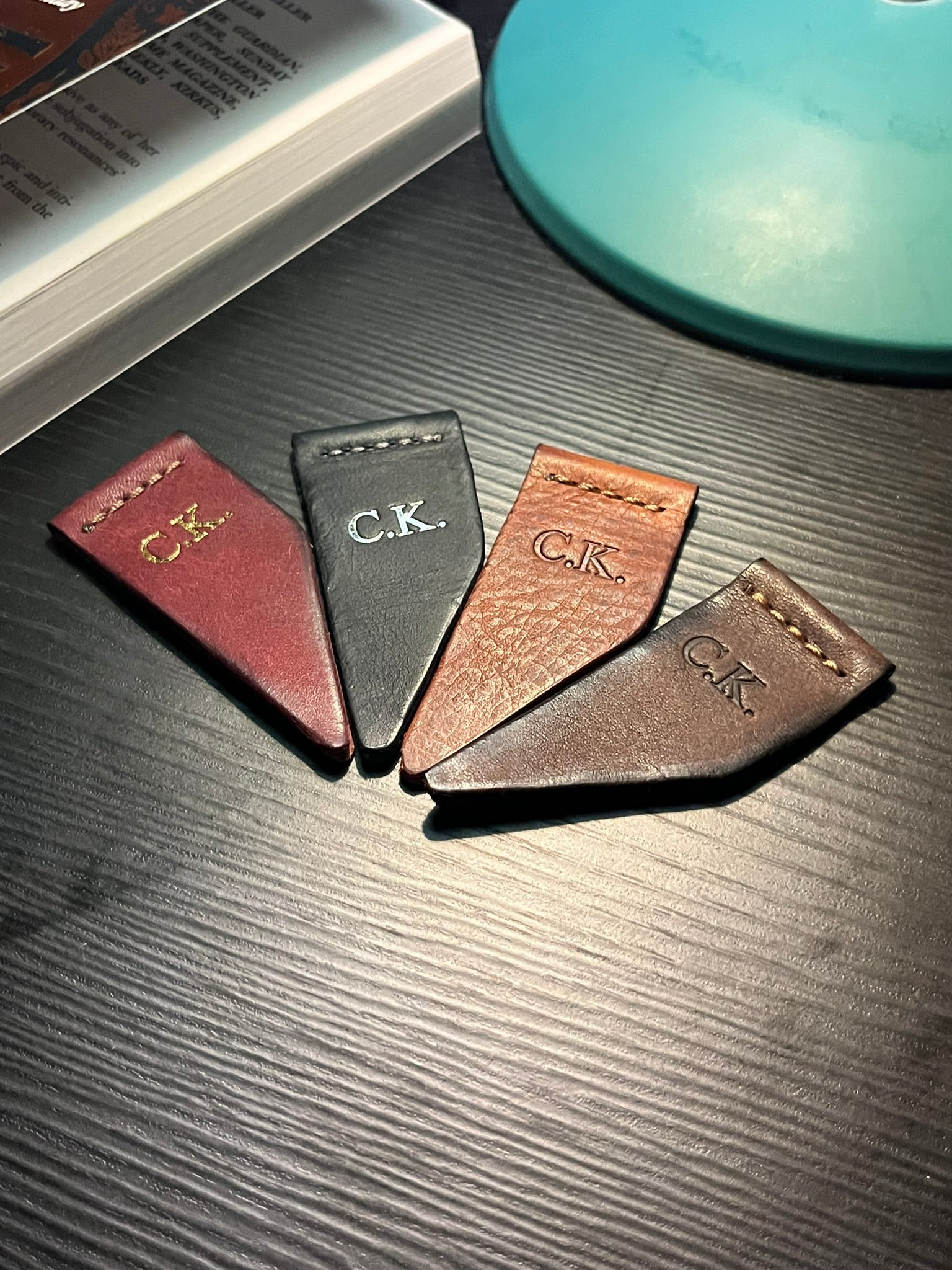Four leather bookmarks with initials 'C.K.' foiling and debossing laying on a dark surface.