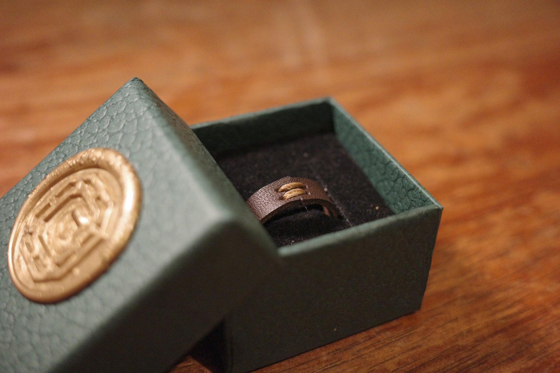 Green jewelry box with a gold emblem on a wooden surface with a brown leather ring inside